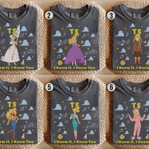 May include: Six charcoal gray t-shirts featuring different illustrated characters, the letters TS, musical notes, clouds, stars, and the text I KNEW IT, I KNEW YOU. Each shirt displays a unique character in various outfits including dresses and jumpsuits.