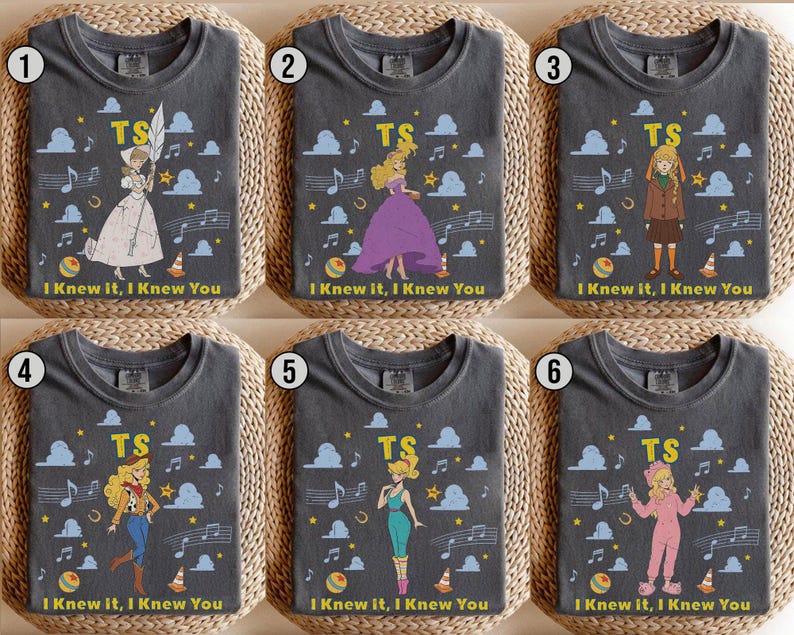 May include: Six charcoal gray t-shirts featuring different illustrated characters, the letters TS, musical notes, clouds, stars, and the text I KNEW IT, I KNEW YOU. Each shirt displays a unique character in various outfits including dresses and jumpsuits.