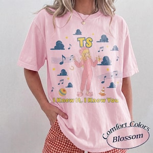 May include: A light pink short-sleeve t-shirt featuring a graphic of a blonde character in a pink onesie, surrounded by blue clouds, musical notes, stars, a ball, and a traffic cone. The text TS and I Knew It, I Knew You are printed in yellow.