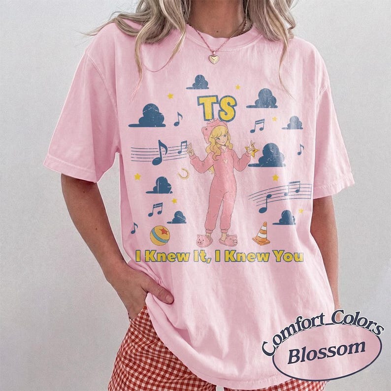 May include: A light pink short-sleeve t-shirt featuring a graphic of a blonde character in a pink onesie, surrounded by blue clouds, musical notes, stars, a ball, and a traffic cone. The text TS and I Knew It, I Knew You are printed in yellow.