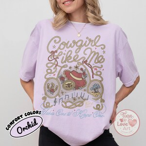 Pu&ograve; includere: Una t-shirt color orchidea con una stampa grafica che riporta il testo "Cowgirl Like Me" e "Takes One to Know One". Il design include bordi a corda, un cappello da cowboy rosso, un ferro di cavallo, un paesaggio desertico e note musicali.