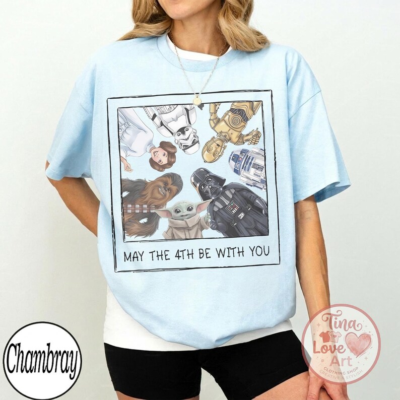 May include: Light blue t-shirt featuring a framed graphic of Star Wars characters including Darth Vader, Yoda, and Chewbacca. The text "MAY THE 4TH BE WITH YOU" is below the graphic. The shirt is a light blue color.