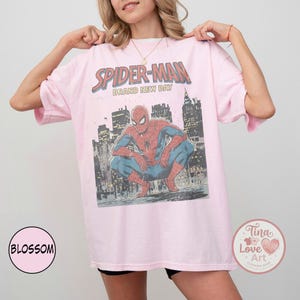 May include: A light pink Spider-Man graphic t-shirt. The design features Spider-Man crouching in front of a city skyline, with the words "SPIDER-MAN BRAND NEW DAY" above. The shirt is a relaxed fit.