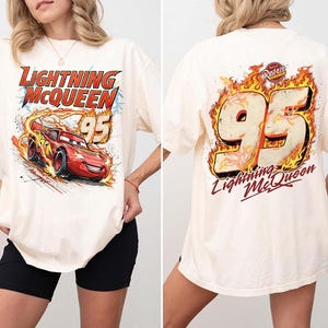 May include: An off-white short-sleeve t-shirt featuring a graphic of the red race car Lightning McQueen with the number 95 on the front. The back of the shirt displays a large number 95 surrounded by flames and the text Lightning McQueen.