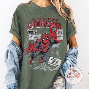 Może przedstawiać: Oliwkowo-zielona koszulka z nadrukiem w stylu vintage komiksu Spider-Man. Grafika zawiera tekst "the AMAZING SPIDER-MAN" i "MARVEL COMICS GROUP". Projekt przedstawia Spider-Mana w dynamicznej pozie z wyciągniętymi rękami.