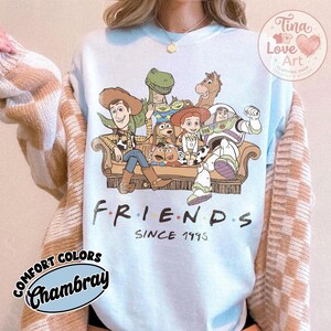 Peut inclure: Un sweat-shirt bleu clair &agrave; col rond orn&eacute; d'une illustration de personnages de Toy Story assis sur un canap&eacute;, avec le texte "FRIENDS SINCE 1995" imprim&eacute; en dessous. Les personnages incluent Woody, Buzz l'&Eacute;clair, Jessie, Rex et Bullseye.