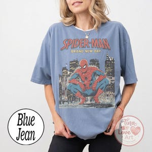 May include: A blue t-shirt featuring a graphic of Spider-Man in a crouching pose against a city skyline. The text "SPIDER-MAN BRAND NEW DAY" is above the image. The shirt has short sleeves and a round neckline.