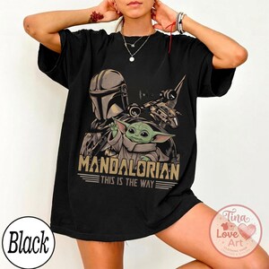 Pu&ograve; includere: T-shirt nera con grafica di The Mandalorian e Grogu, con la scritta "MANDALORIAN THIS IS THE WAY". Il design include un casco, un'astronave e un personaggio alieno verde. La maglietta ha scollo tondo e maniche corte.