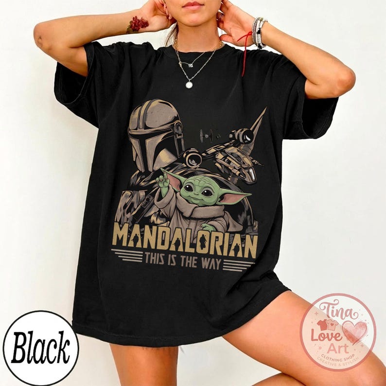 Pu&ograve; includere: T-shirt nera con grafica di The Mandalorian e Grogu, con la scritta "MANDALORIAN THIS IS THE WAY". Il design include un casco, un'astronave e un personaggio alieno verde. La maglietta ha scollo tondo e maniche corte.