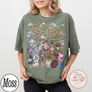 Op de afbeelding: Een olijfgroen T-shirt met korte mouwen en een grafische print van diverse geanimeerde personages uit de Toy Story-franchise. Het ontwerp bevat de tekst Toy Story in een touwachtig lettertype, samen met een ster, wolken en speelgoedfiguren.