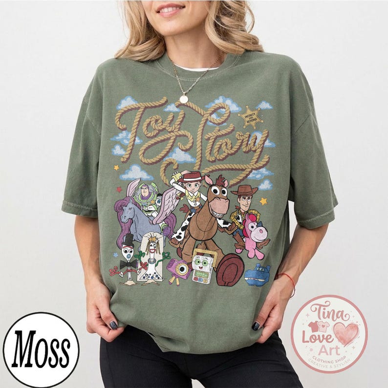 Op de afbeelding: Een olijfgroen T-shirt met korte mouwen en een grafische print van diverse geanimeerde personages uit de Toy Story-franchise. Het ontwerp bevat de tekst Toy Story in een touwachtig lettertype, samen met een ster, wolken en speelgoedfiguren.