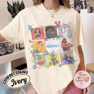 Puede incluir: Camiseta de color marfil con una cuadr&iacute;cula de ilustraciones coloridas de personajes de la pel&iacute;cula de Disney Moana. La camiseta tiene las palabras "Comfort Colors" e "Ivory" impresas. Es visible una pegatina redonda con el texto "Tina Love Art".