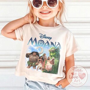 May include: A light beige t-shirt featuring the Disney movie "Moana" logo in blue and a graphic of the characters. The shirt is short-sleeved and has a crew neck. A small, round logo is visible on the lower right.