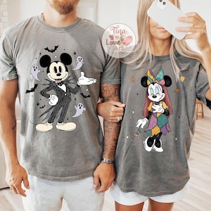 Op de afbeelding: Twee grijze t-shirts met Disney-personages met Halloween-thema. E&eacute;n shirt toont Mickey Mouse verkleed als Jack Skellington, omringd door vleermuizen en geesten. Het andere shirt toont Minnie Mouse in een patchwork jurk met herfstbladeren.