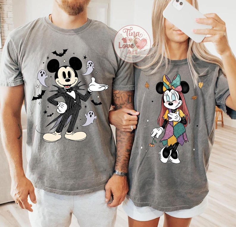 Op de afbeelding: Twee grijze t-shirts met Disney-personages met Halloween-thema. E&eacute;n shirt toont Mickey Mouse verkleed als Jack Skellington, omringd door vleermuizen en geesten. Het andere shirt toont Minnie Mouse in een patchwork jurk met herfstbladeren.