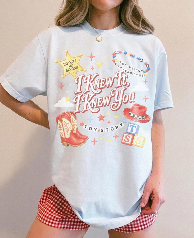 Puede incluir: Una camiseta azul claro con un estampado gr&aacute;fico que incluye el texto "I KNEW IT, I KNEW YOU" y "TOY STORY". El dise&ntilde;o presenta una estrella de sheriff amarilla, un collar de cuentas, botas de vaquero rojas, un sombrero rojo y bloques.