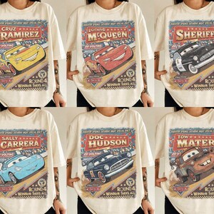 May include: A cream-coloured t-shirt featuring a vintage-style graphic print of Disney Pixar Cars characters including Cruz Ramirez, Lightning McQueen, Sheriff, Sally Carrera, Doc Hudson, and Tow Mater with the text Radiator Springs Saturday Night Special Race.