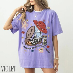 May include: A light purple short-sleeve t-shirt featuring a graphic print of a cowboy boot with a cow print pattern, a red hat, and stacked blocks with the letters T, S, and 13. The text Taylor is on the boot, and a beaded chain design surrounds it.