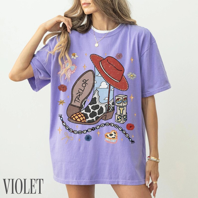 May include: A light purple short-sleeve t-shirt featuring a graphic print of a cowboy boot with a cow print pattern, a red hat, and stacked blocks with the letters T, S, and 13. The text Taylor is on the boot, and a beaded chain design surrounds it.