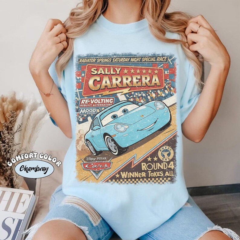 May include: A light blue t-shirt featuring a graphic print of the character Sally Carrera from the movie Cars. The design includes the text Radiator Springs Saturday Night Special Race, Sally Carrera, Re-Volting, Mood Springs, and Round 4 Winner Takes All.