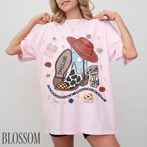 May include: A light pink t-shirt featuring a graphic print of a cowboy boot with a cow print pattern, a red cowboy hat, a blue sky background, the name TAYLOR, the number 13, alphabet blocks spelling TS, a bow, and a beaded string with text.