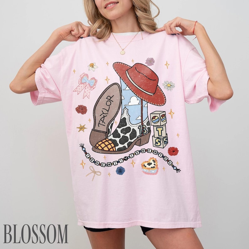 May include: A light pink t-shirt featuring a graphic print of a cowboy boot with a cow print pattern, a red cowboy hat, a blue sky background, the name TAYLOR, the number 13, alphabet blocks spelling TS, a bow, and a beaded string with text.