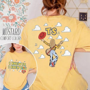 K&ouml;nnte beinhalten: Ein senfgelbes T-Shirt mit einer Grafik auf der R&uuml;ckseite, die eine Person mit einer Gitarre und einem roten Hut zeigt, umgeben von Wolken und den Buchstaben TS. Die Vorderseite zeigt den Text I KNEW IT, I KNEW YOU mit einem Herz und TS.