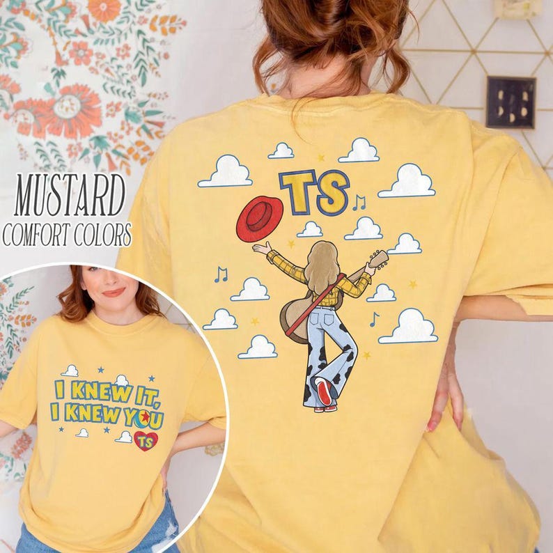 K&ouml;nnte beinhalten: Ein senfgelbes T-Shirt mit einer Grafik auf der R&uuml;ckseite, die eine Person mit einer Gitarre und einem roten Hut zeigt, umgeben von Wolken und den Buchstaben TS. Die Vorderseite zeigt den Text I KNEW IT, I KNEW YOU mit einem Herz und TS.