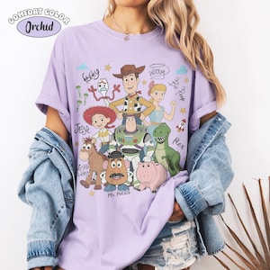 May include: A light purple t-shirt featuring a graphic print of Toy Story characters including Woody, Buzz Lightyear, Jessie, Rex, Mr. Potato Head, Bullseye, Bo Peep, and Forky, with handwritten-style character names and small star and cloud icons.