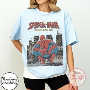 May include: Light blue t-shirt featuring a graphic of Spider-Man in a crouching pose against a cityscape backdrop. The words "SPIDER-MAN" and "BRAND NEW DAY" are printed above the image in red and white.