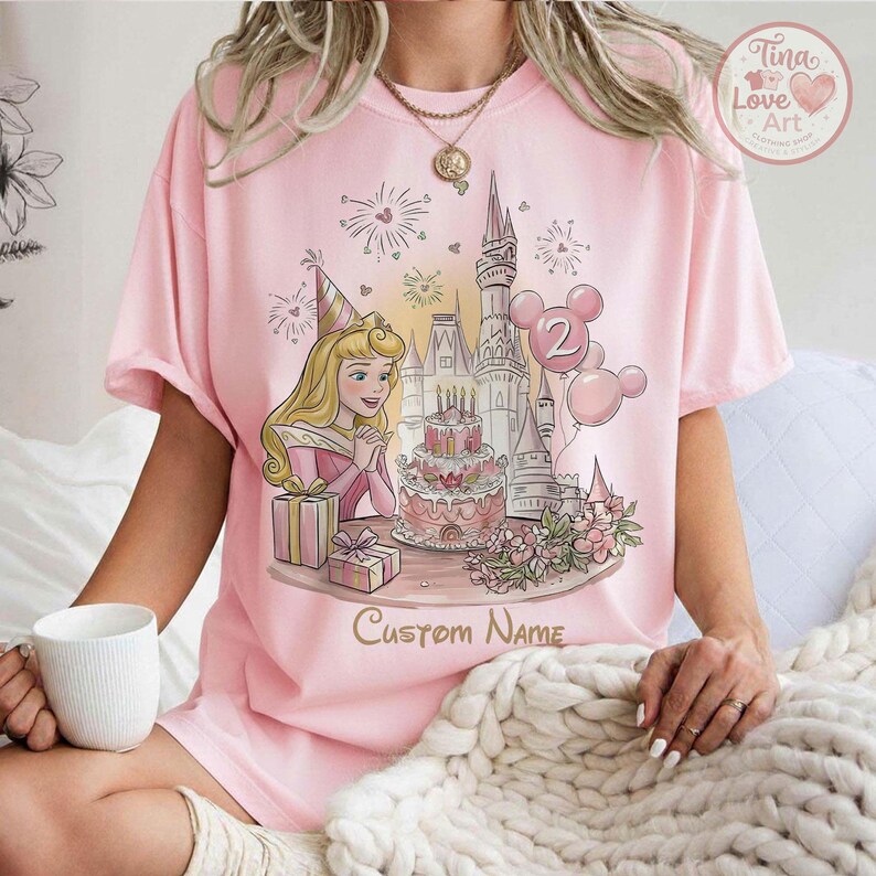 May include: Pale pink t-shirt with a princess birthday design. The illustration shows a castle, cake, balloons, and the words "Custom Name". The design uses soft pastel colours, with the princess wearing a party hat.