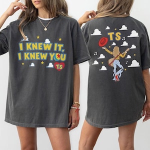 K&ouml;nnte beinhalten: Ein anthrazitfarbenes T-Shirt mit einem Frontaufdruck mit dem gelben Text I KNEW IT, I KNEW YOU und einem roten Herz mit TS. Die R&uuml;ckseite zeigt eine Person mit langem Haar, die eine Gitarre und einen roten Hut h&auml;lt, umgeben von wei&szlig;en Wolken.