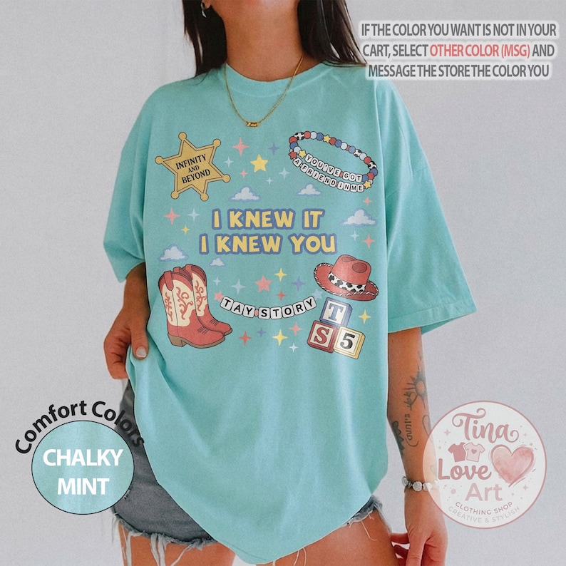 May include: A light mint green t-shirt featuring a graphic print with a yellow sheriff star, a beaded bracelet, a red cowboy boot, and alphabet blocks. The text reads: "INFINITY AND BEYOND", "YOU'VE GOT A FRIEND IN ME", "I KNEW IT I KNEW YOU", and "TAY STORY".