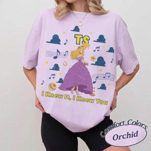 May include: A light purple t-shirt featuring a graphic of a blonde character in a purple gown, surrounded by blue clouds, musical notes, stars, and a ball. The text TS and I KNEW IT, I KNEW YOU are printed in yellow with a dark outline on the front.