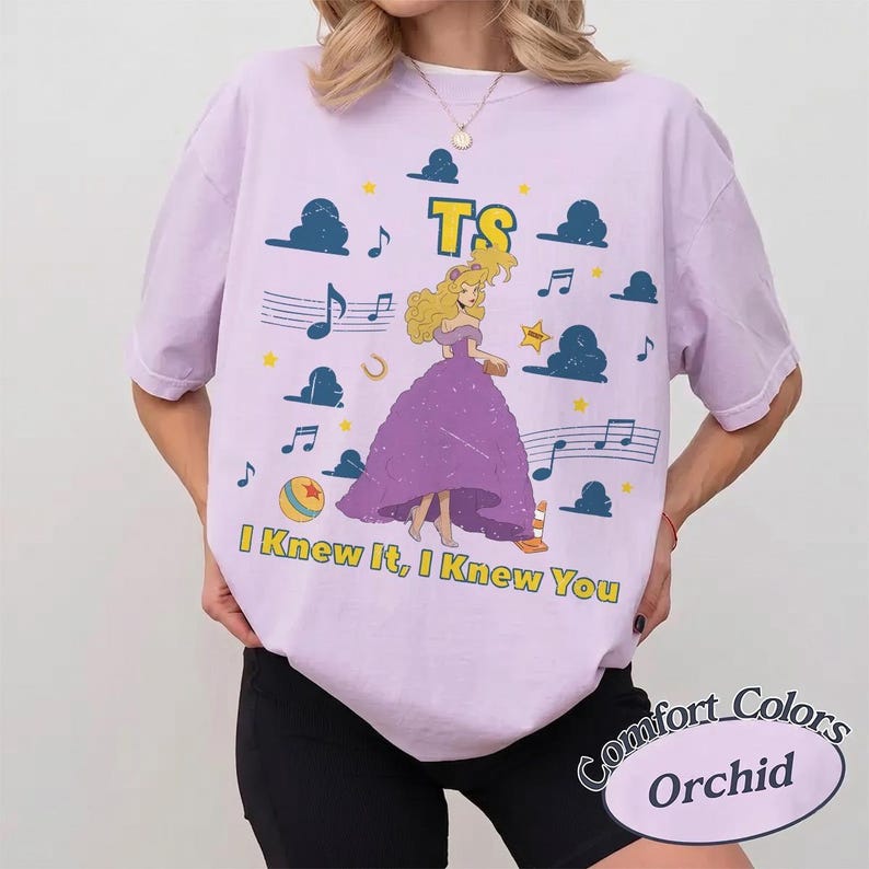 May include: A light purple t-shirt featuring a graphic of a blonde character in a purple gown, surrounded by blue clouds, musical notes, stars, and a ball. The text TS and I KNEW IT, I KNEW YOU are printed in yellow with a dark outline on the front.