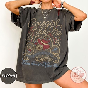 Pu&ograve; includere: Una t-shirt grigio antracite a maniche corte con una stampa grafica che riporta il testo Cowgirl like Me e Takes One to Know One. Il design include un cappello da cowboy rosso, bordi a corda, rose, un ferro di cavallo e note musicali.