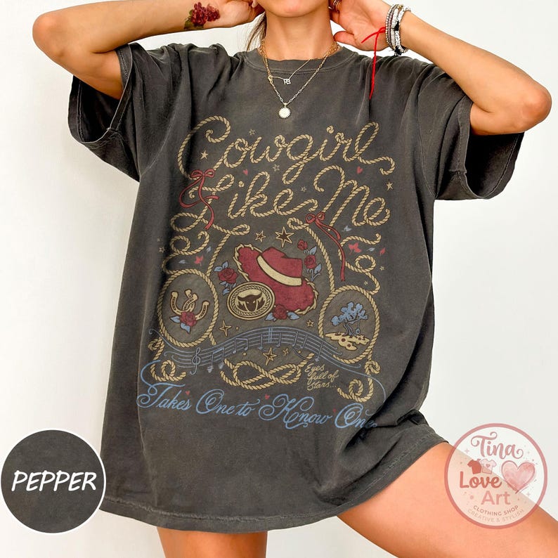 Pu&ograve; includere: Una t-shirt grigio antracite a maniche corte con una stampa grafica che riporta il testo Cowgirl like Me e Takes One to Know One. Il design include un cappello da cowboy rosso, bordi a corda, rose, un ferro di cavallo e note musicali.