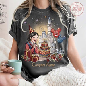May include: Dark grey t-shirt with a cartoon illustration of a woman in a red and blue outfit, a birthday cake, and a castle. The image includes the text "Custom Name" and balloons with the number "2".