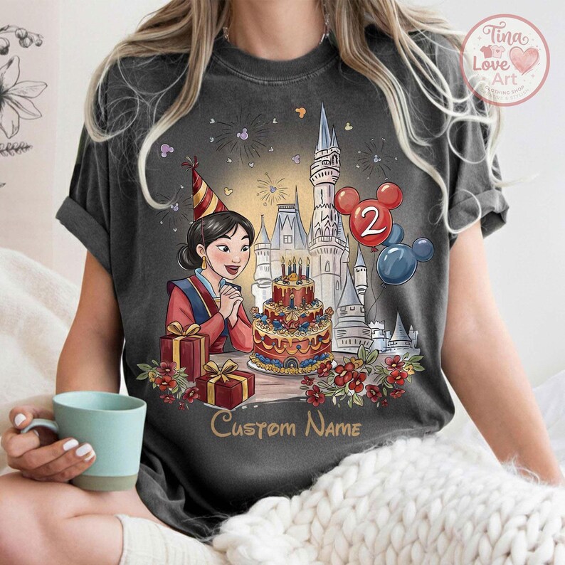 May include: Dark grey t-shirt with a cartoon illustration of a woman in a red and blue outfit, a birthday cake, and a castle. The image includes the text "Custom Name" and balloons with the number "2".