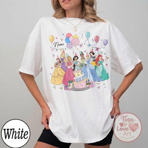 May include: White t-shirt featuring a colorful illustration of several princesses around a birthday cake. The design includes balloons, the number 5, and the word "Name". The shirt has short sleeves and a relaxed fit.