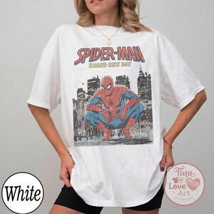 May include: White t-shirt featuring a Spider-Man graphic. The design includes the text "SPIDER-MAN BRAND NEW DAY" above an illustration of Spider-Man crouching in front of a cityscape. The shirt is a casual, oversized fit.