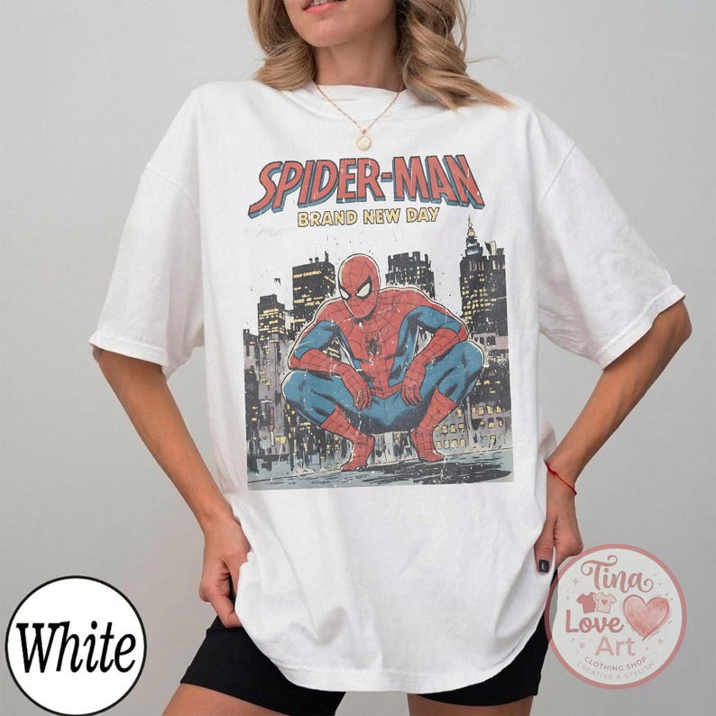 May include: White t-shirt featuring a Spider-Man graphic. The design includes the text "SPIDER-MAN BRAND NEW DAY" above an illustration of Spider-Man crouching in front of a cityscape. The shirt is a casual, oversized fit.