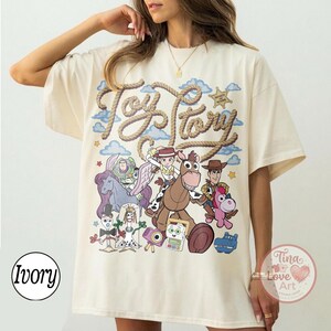 Op de afbeelding: Een ivoorkleurig t-shirt met een grote grafische print met de tekst Toy Story in een touwachtig lettertype. Het ontwerp bevat kleurrijke illustraties van verschillende geanimeerde personages, waaronder een space ranger, een cowgirl, een cowboy en een paard.