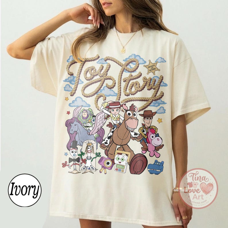 Op de afbeelding: Een ivoorkleurig t-shirt met een grote grafische print met de tekst Toy Story in een touwachtig lettertype. Het ontwerp bevat kleurrijke illustraties van verschillende geanimeerde personages, waaronder een space ranger, een cowgirl, een cowboy en een paard.
