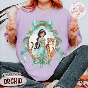 May include: A lilac Comfort Color t-shirt featuring a framed illustration of Princess Jasmine and a tiger. The frame is adorned with teal ribbons and bows. The text "Comfort Color" and "Orchid" are also visible.
