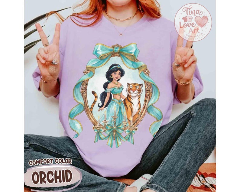May include: A lilac Comfort Color t-shirt featuring a framed illustration of Princess Jasmine and a tiger. The frame is adorned with teal ribbons and bows. The text "Comfort Color" and "Orchid" are also visible.