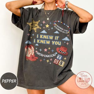 May include: A charcoal gray short-sleeve t-shirt featuring a graphic print with the text I KNEW IT I KNEW YOU, TAY STORY, INFINITY AND BEYOND, YOU'VE GOT A FRIEND IN ME, and TS 5. The design includes red cowboy boots, a red hat, stars, and clouds.