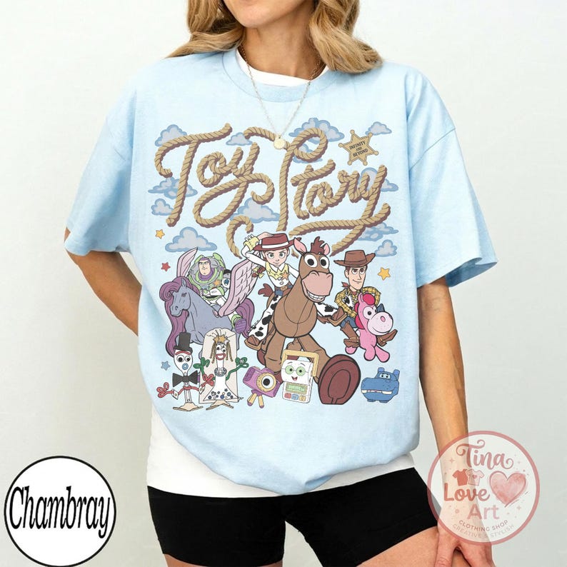 Op de afbeelding: Een lichtblauw t-shirt met een grafische print van Toy Story-personages, waaronder Woody, Jessie, Bullseye, Buzz Lightyear en Forky. De tekst Toy Story staat in een touwachtig lettertype boven de personages tegen een luchtachtergrond.