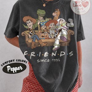 Peut inclure: Un t-shirt gris anthracite &agrave; manches courtes avec une impression graphique de personnages de Toy Story assis sur un canap&eacute;, accompagn&eacute; du texte "F.R.I.E.N.D.S" et "SINCE 1995" imprim&eacute; sous l'illustration. Le t-shirt a un col rond et une coupe d&eacute;contract&eacute;e.