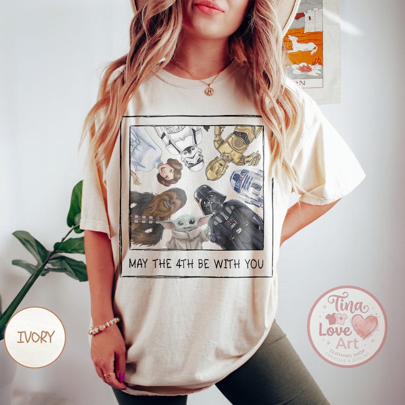 May include: Ivory-colored t-shirt featuring a framed graphic of Star Wars characters including Princess Leia, Darth Vader, and Baby Yoda. The text "MAY THE 4TH BE WITH YOU" is printed below the graphic. The shirt is a comfortable, casual style.
